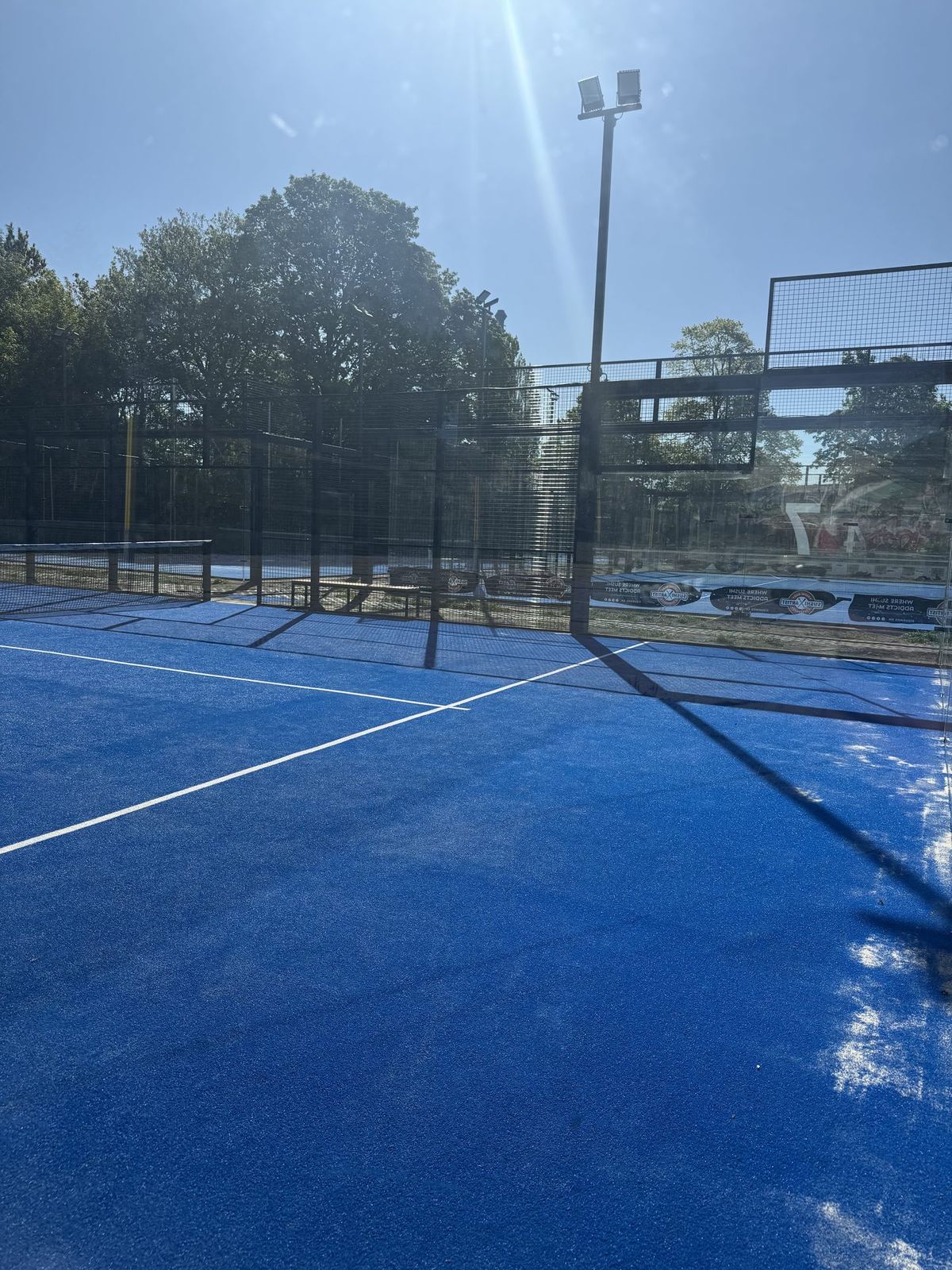 Padel Court mit Sonnenstrahl, blauer Spielfläche und Flutlichtmast